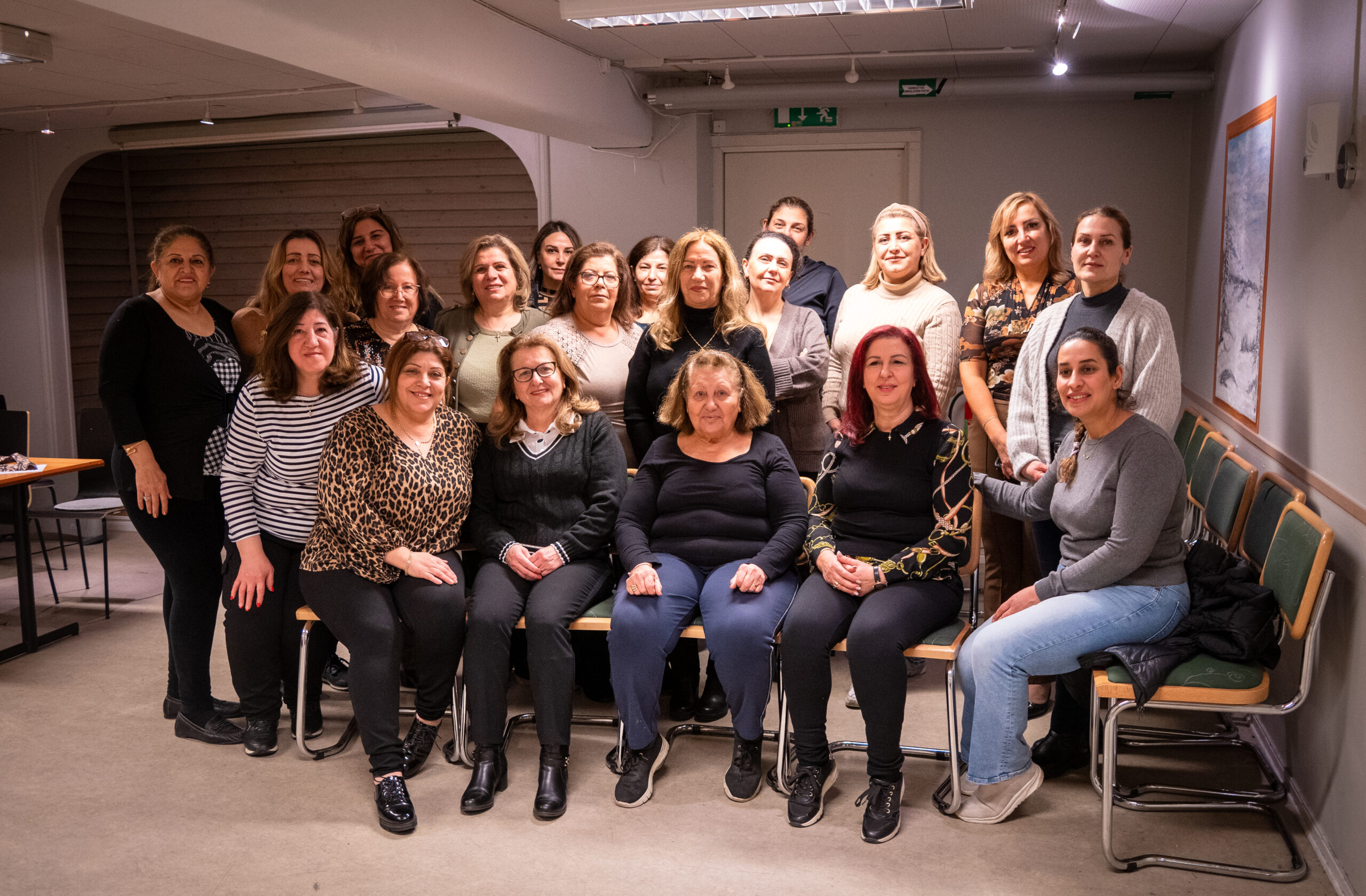 En grupp kvinnor poserar tillsammans inomhus, några sittande på stolar och andra stående bakom dem, leende mot kameran.