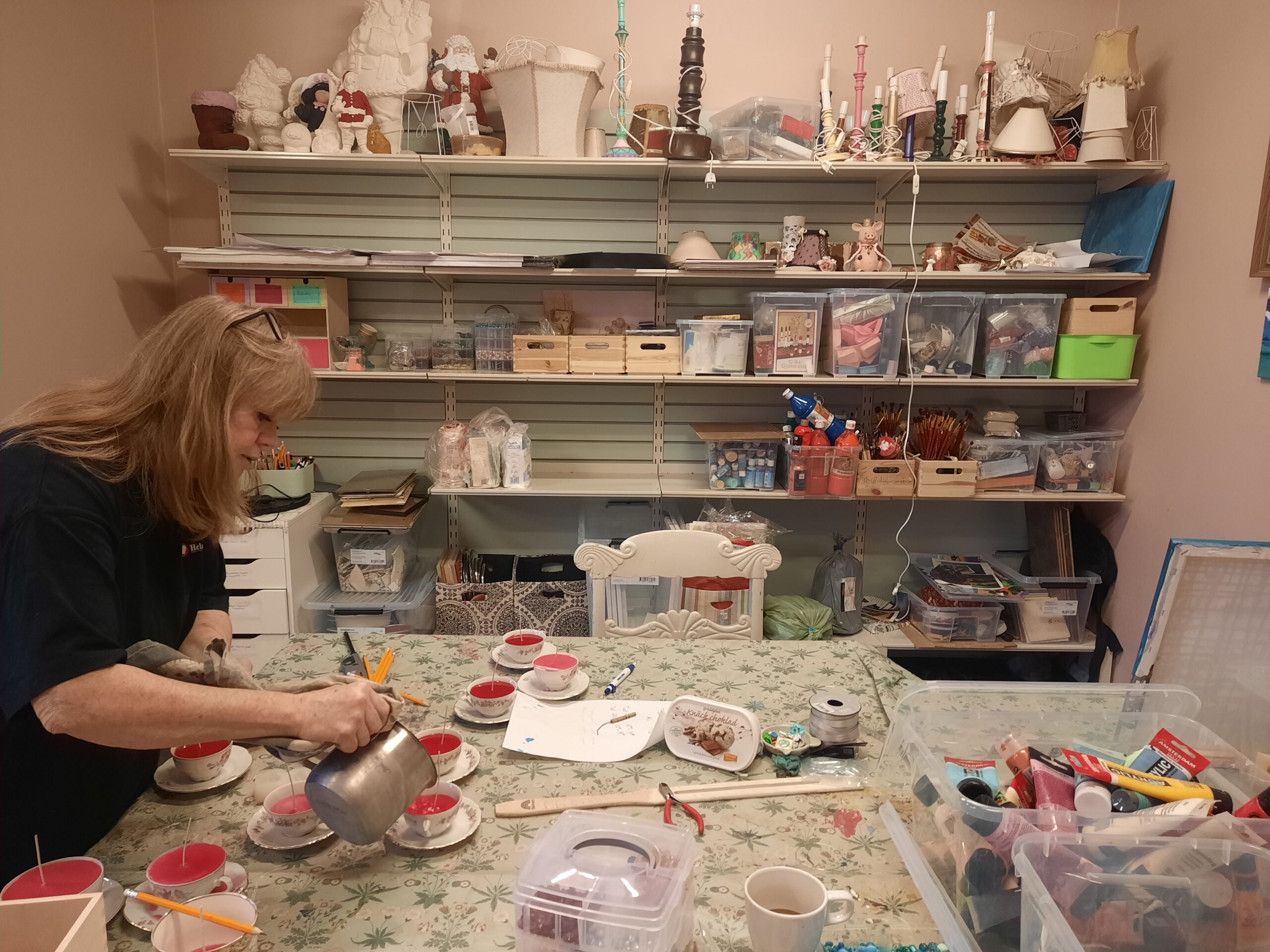 Kvinna häller upp vatten i tekoppar vid ett rörigt pysselbord med hyllor med material i bakgrunden.