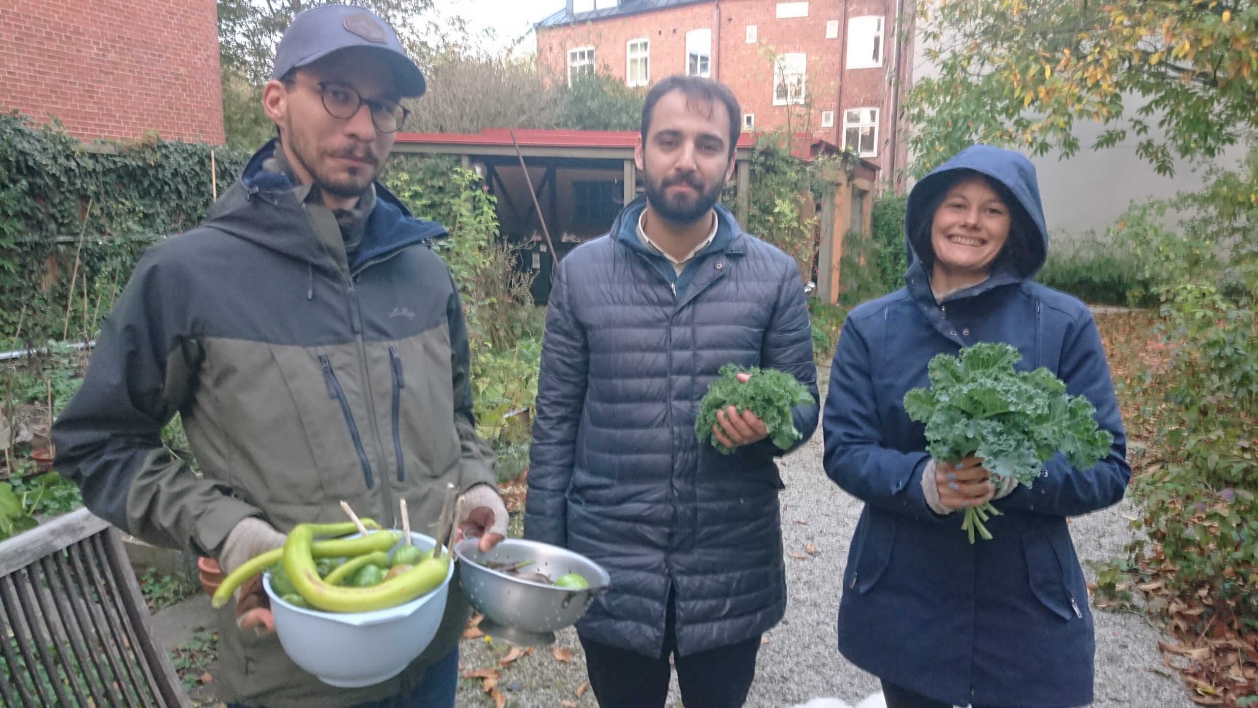 Tre personer i jackor står utanför med nyskördade gröna grönsaker i händerna och ler mot kameran.
