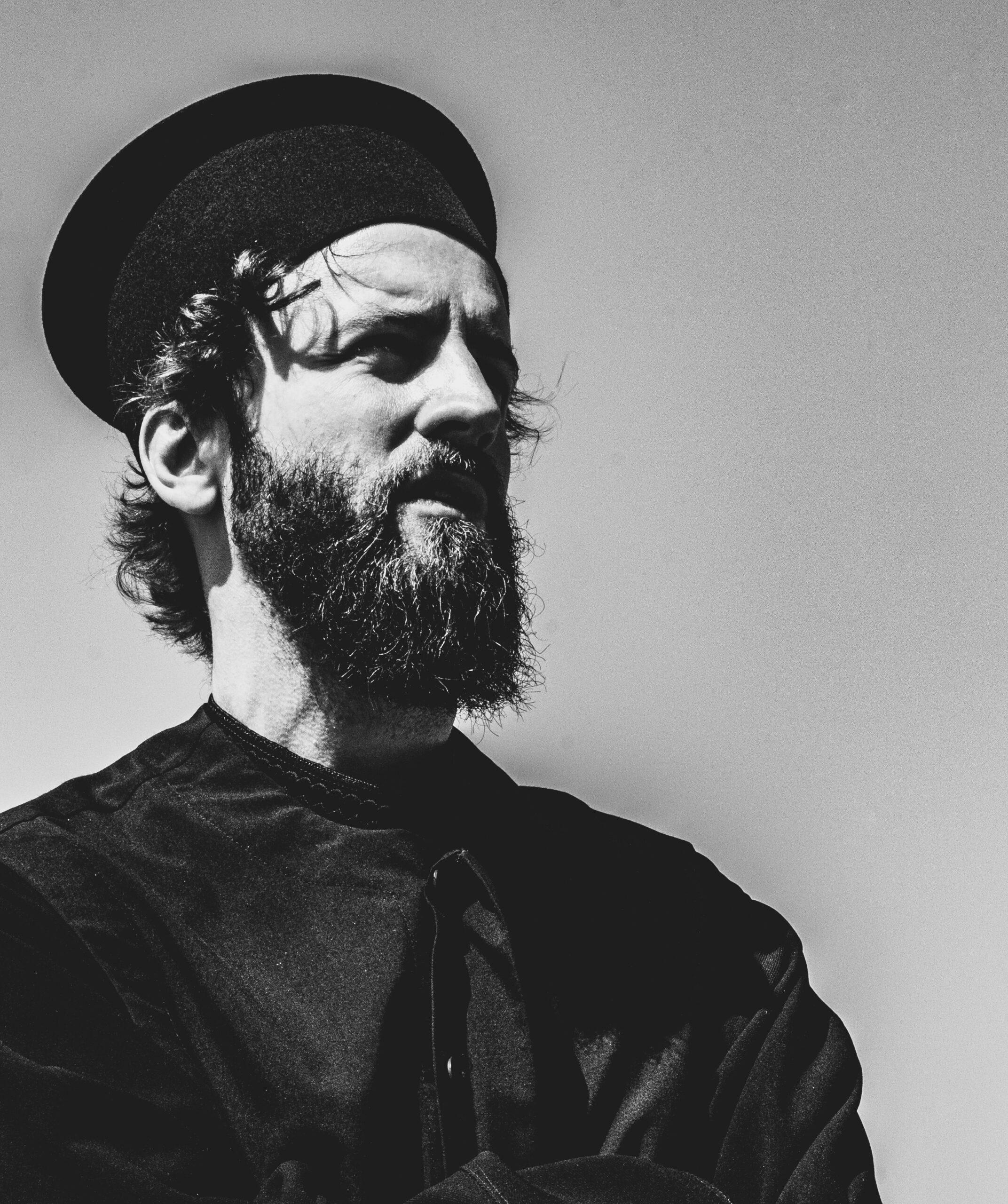 Skäggig man i mörka kläder och rund hatt tittar ut i fjärran mot en enfärgad bakgrund.