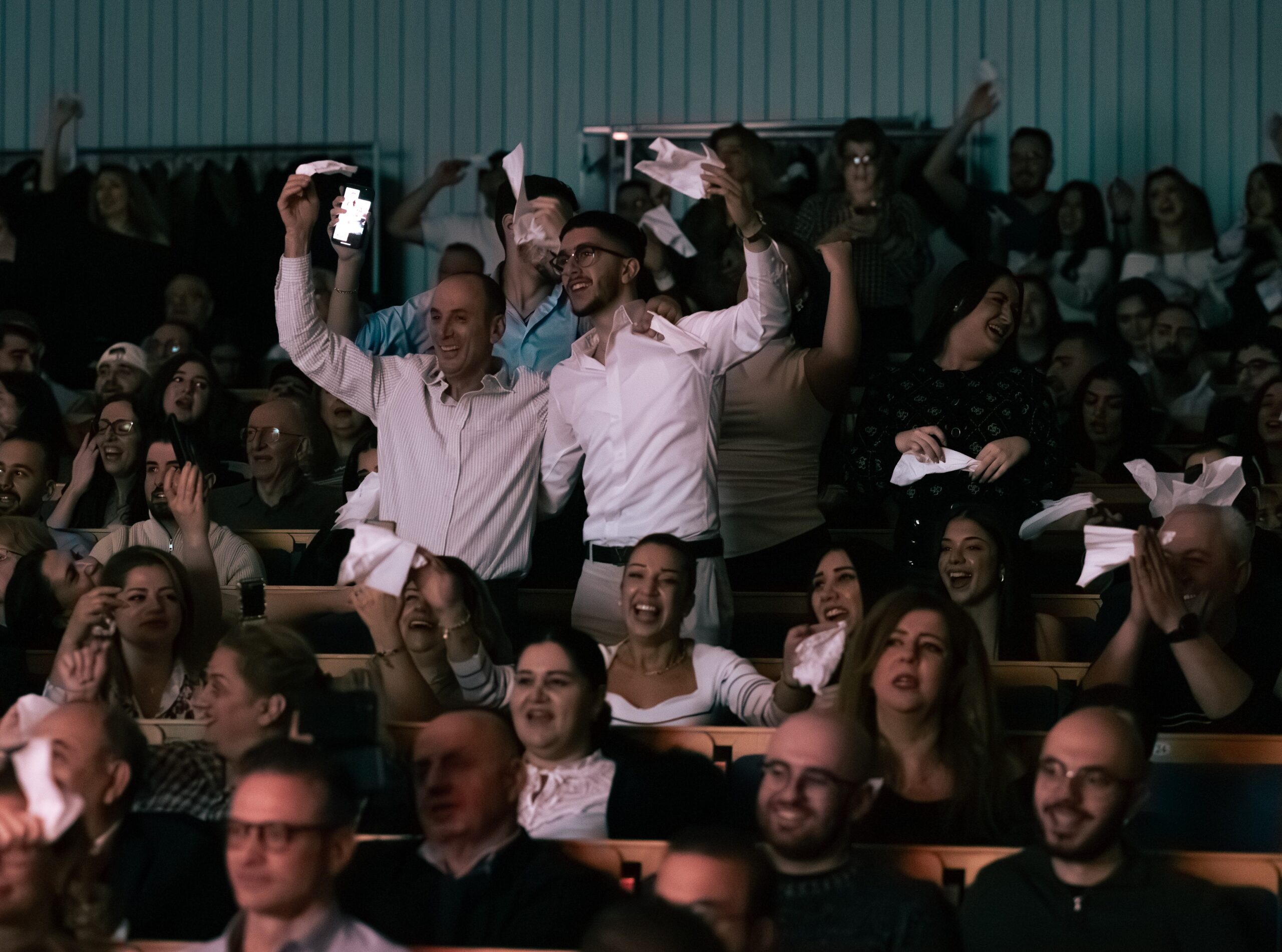 Publiken jublar och viftar med vita servetter vid ett inomhusevenemang, med många människor som ler och applåderar.