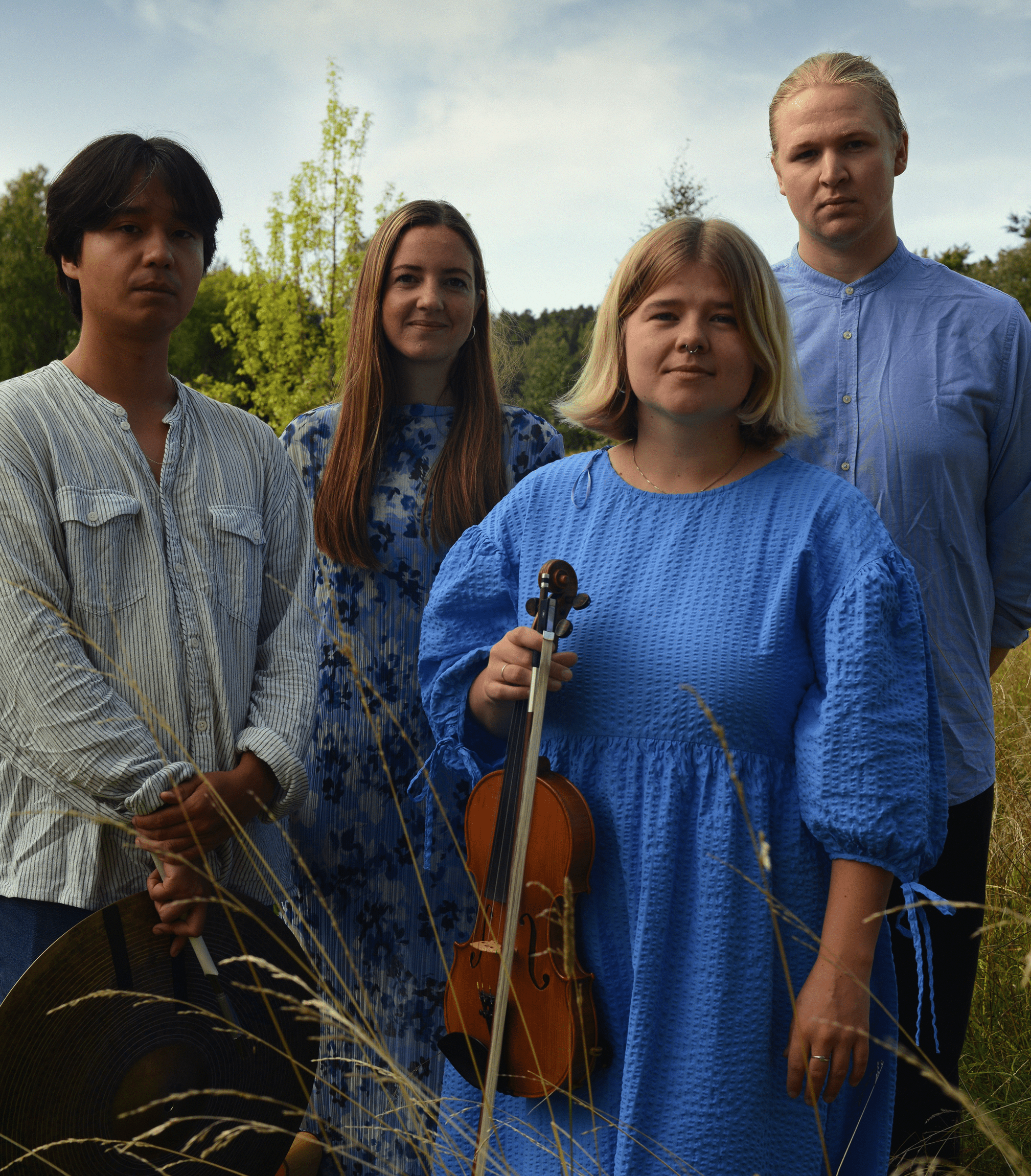 Fyra personer står i högt gräs utomhus; en av dem håller i en fiol. De tittar alla mot kameran.