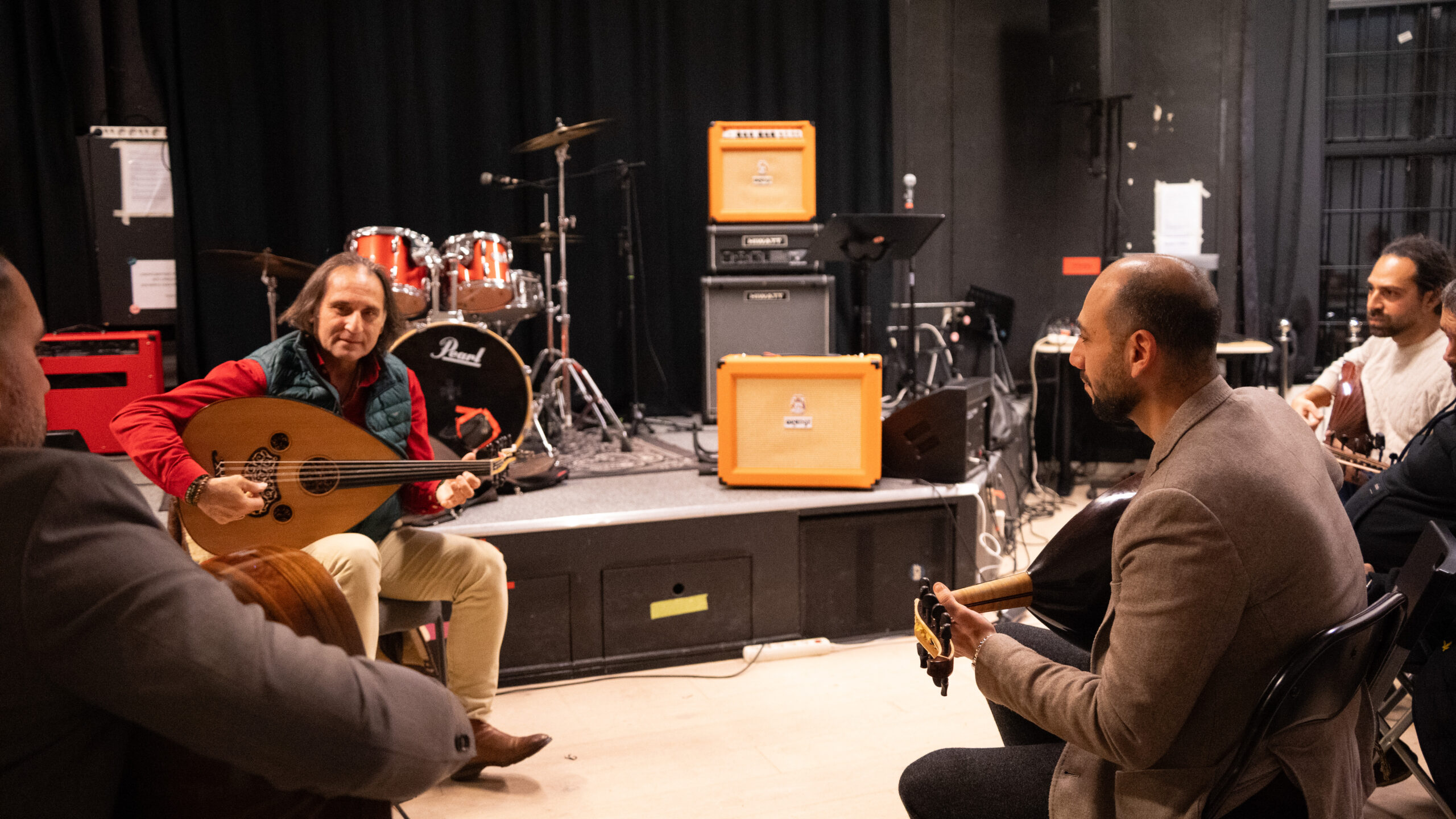 Fyra män spelar stränginstrument i en musikstudio med förstärkare, trummor och svarta gardiner i bakgrunden.
