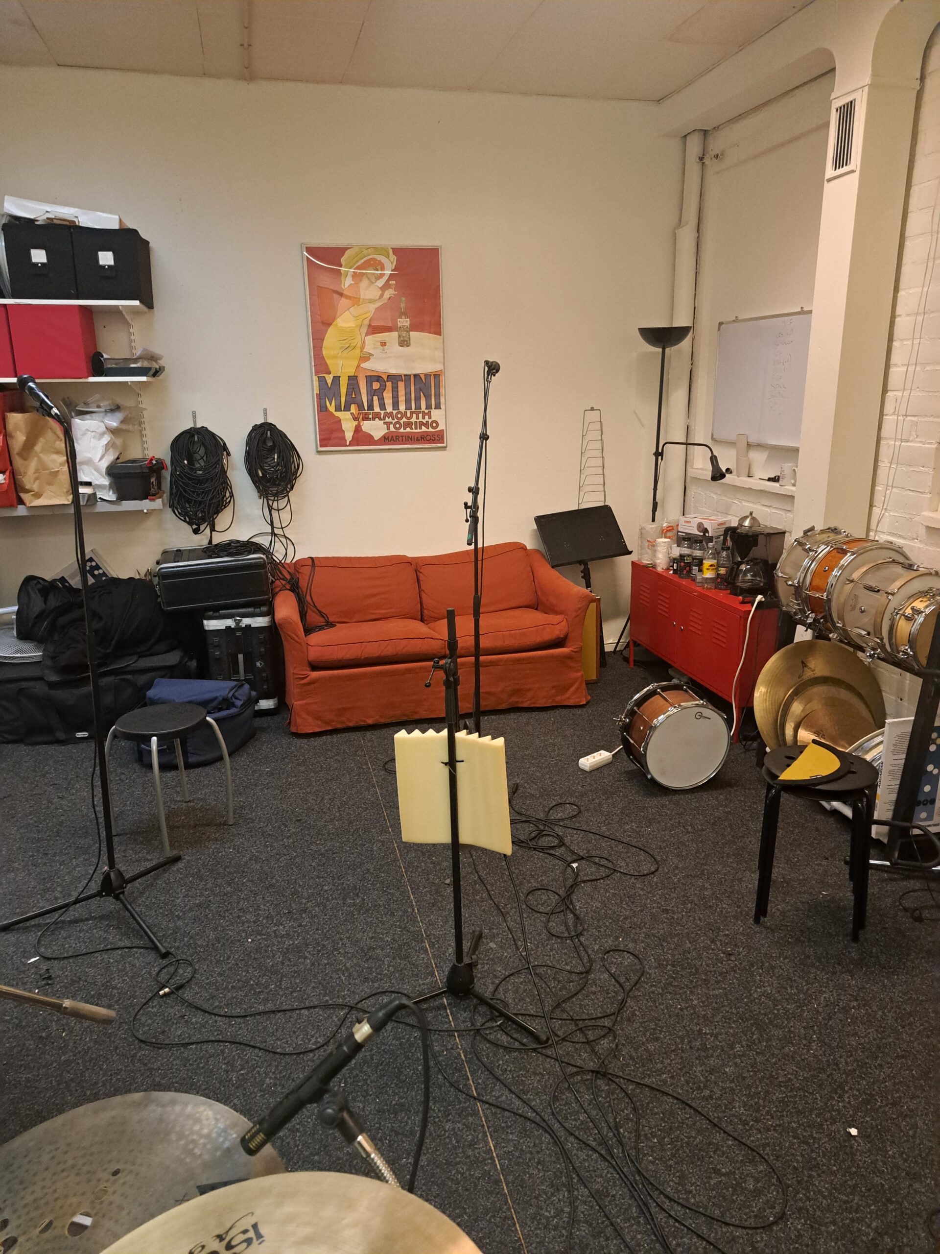 Ett musikrepetitionsrum med en röd soffa, trumset, kablar, mikrofoner och en Martini-affisch på väggen.