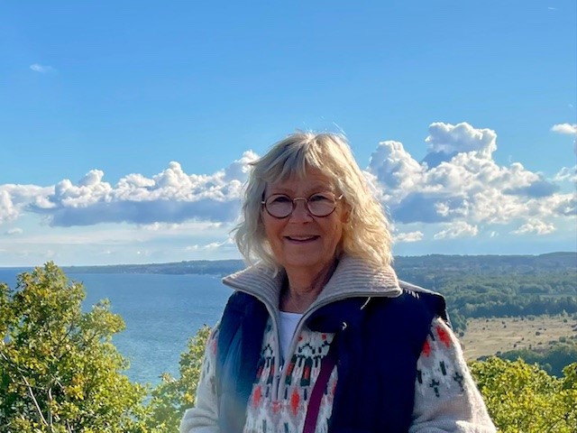 Leende kvinna med glasögon står utomhus vid vatten och träd under en blå himmel med moln.